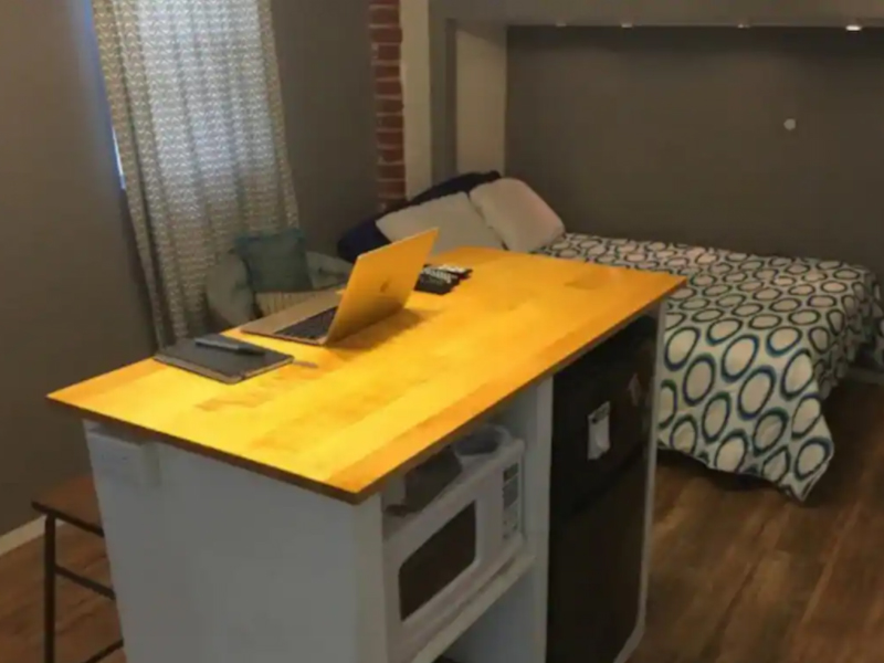 A desk with a laptop and a bed in the background.